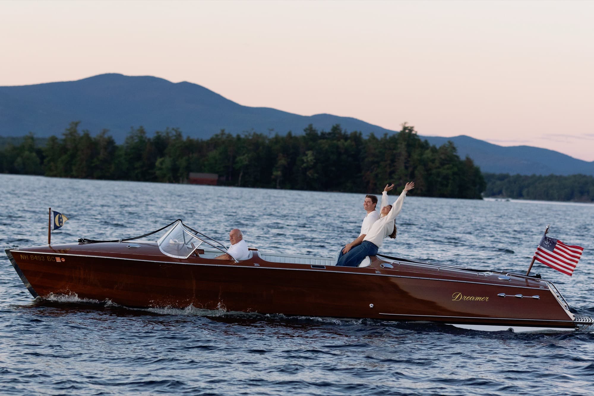 Morgan and Jon in the back of a wooden runabout speeding across the lake at dusk, arms raised, mountains behind them under a pink sky.
