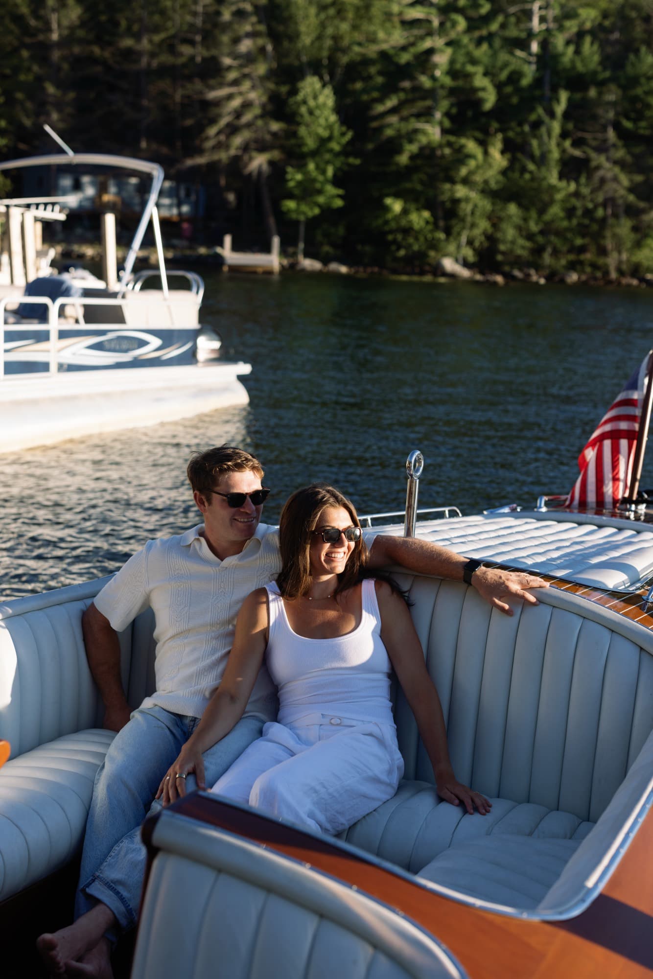 Morgan and Jon relaxed on the rear bench of the boat in sunglasses, shoreline pines across the water.
