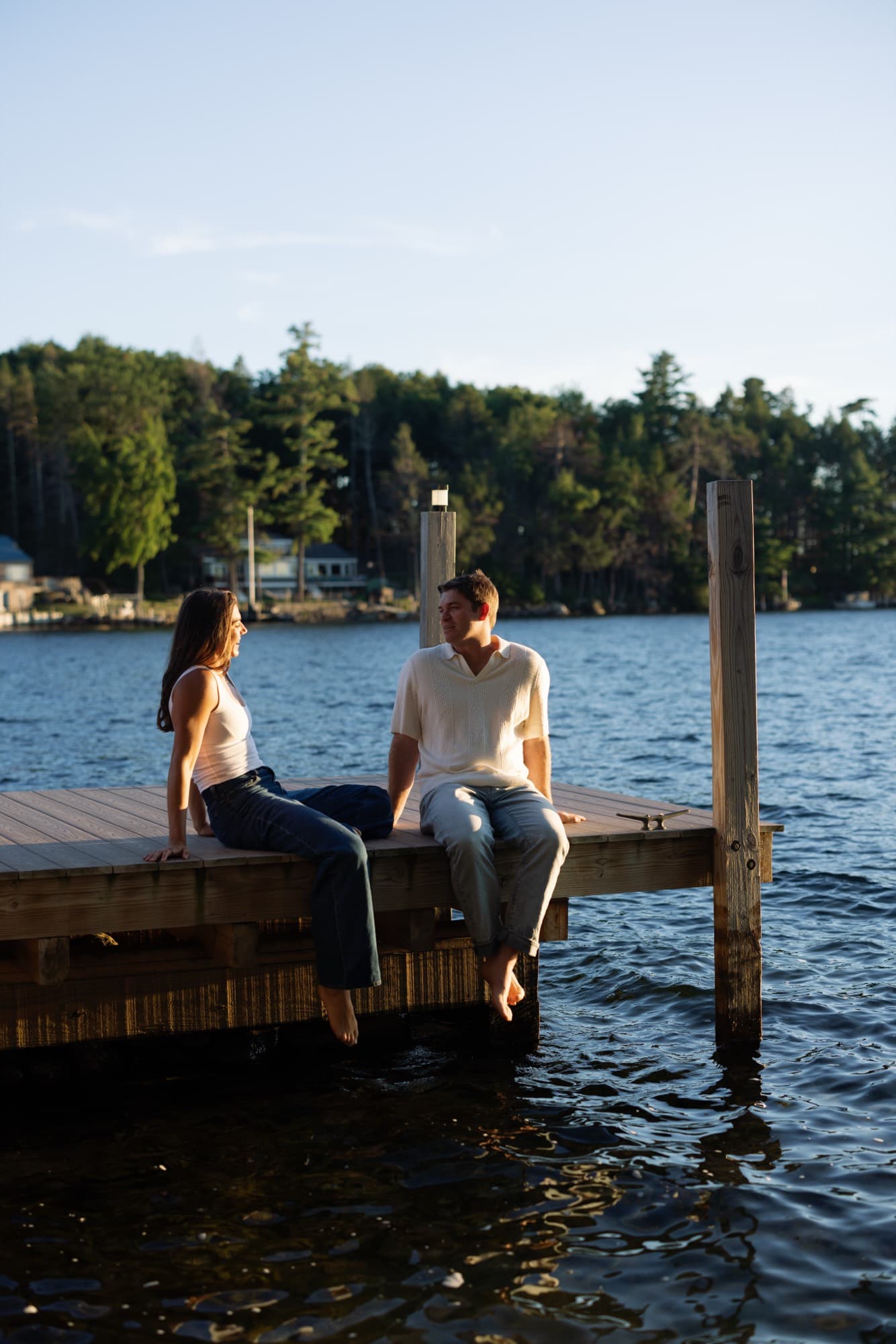 Morgan and Jon sitting on the end of a dock facing each other, bare feet over the water, late afternoon light.
