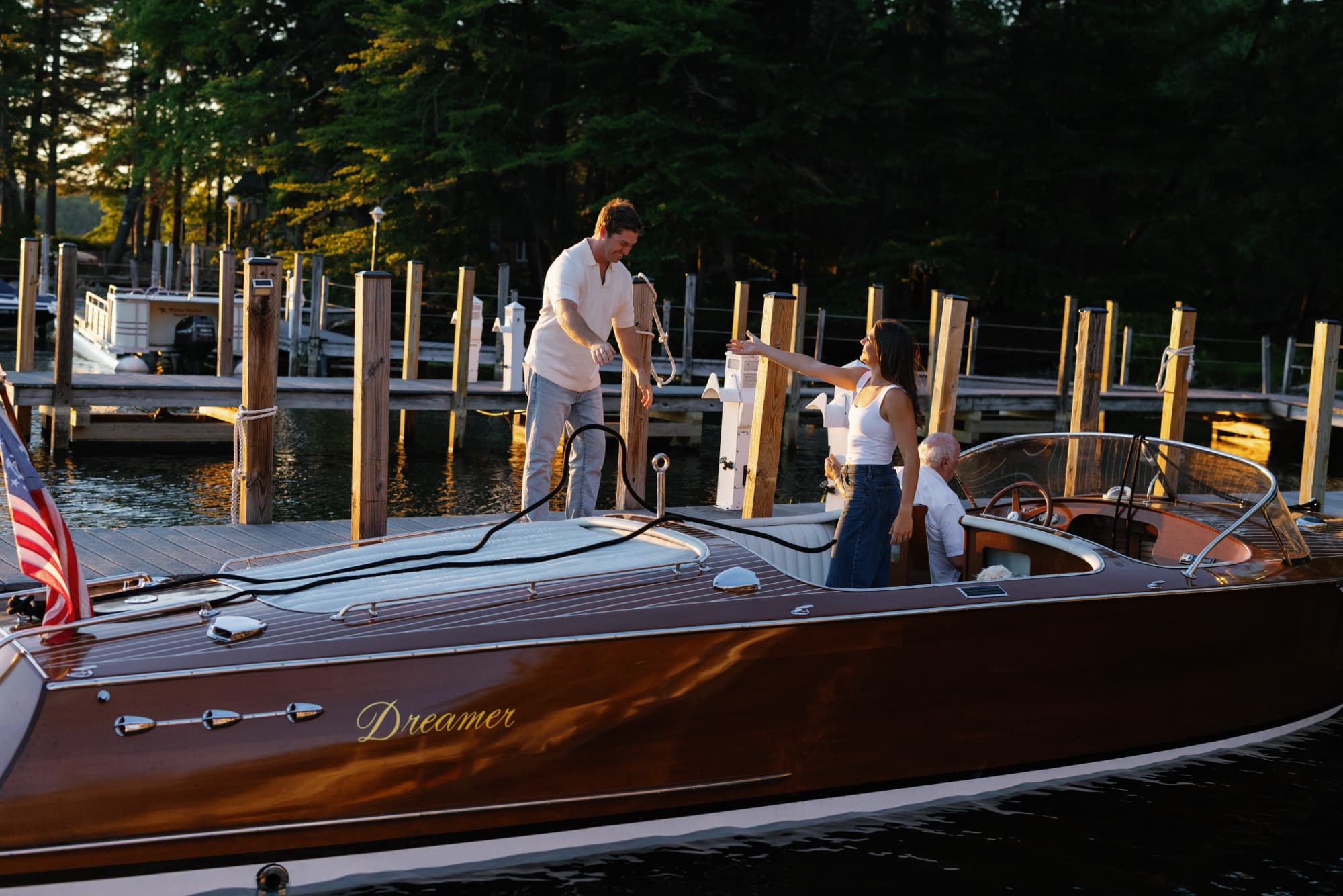 The mahogany runabout pulled alongside a dock at golden hour as Jon hands over the lines and Morgan reaches out from the deck.