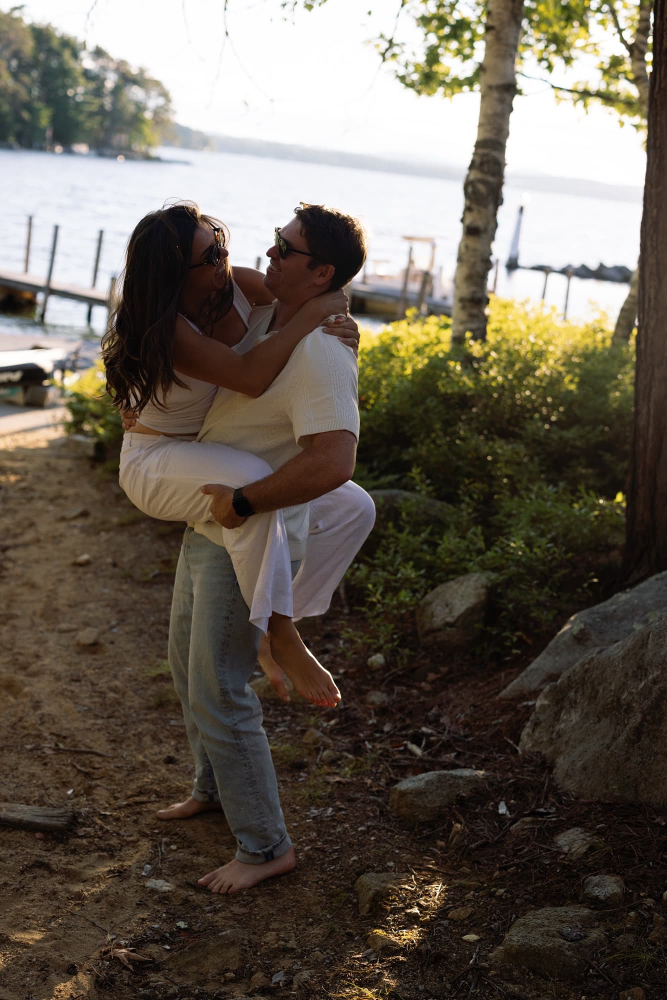 Jon lifting Morgan off her feet on a wooded path by the shoreline, both laughing.