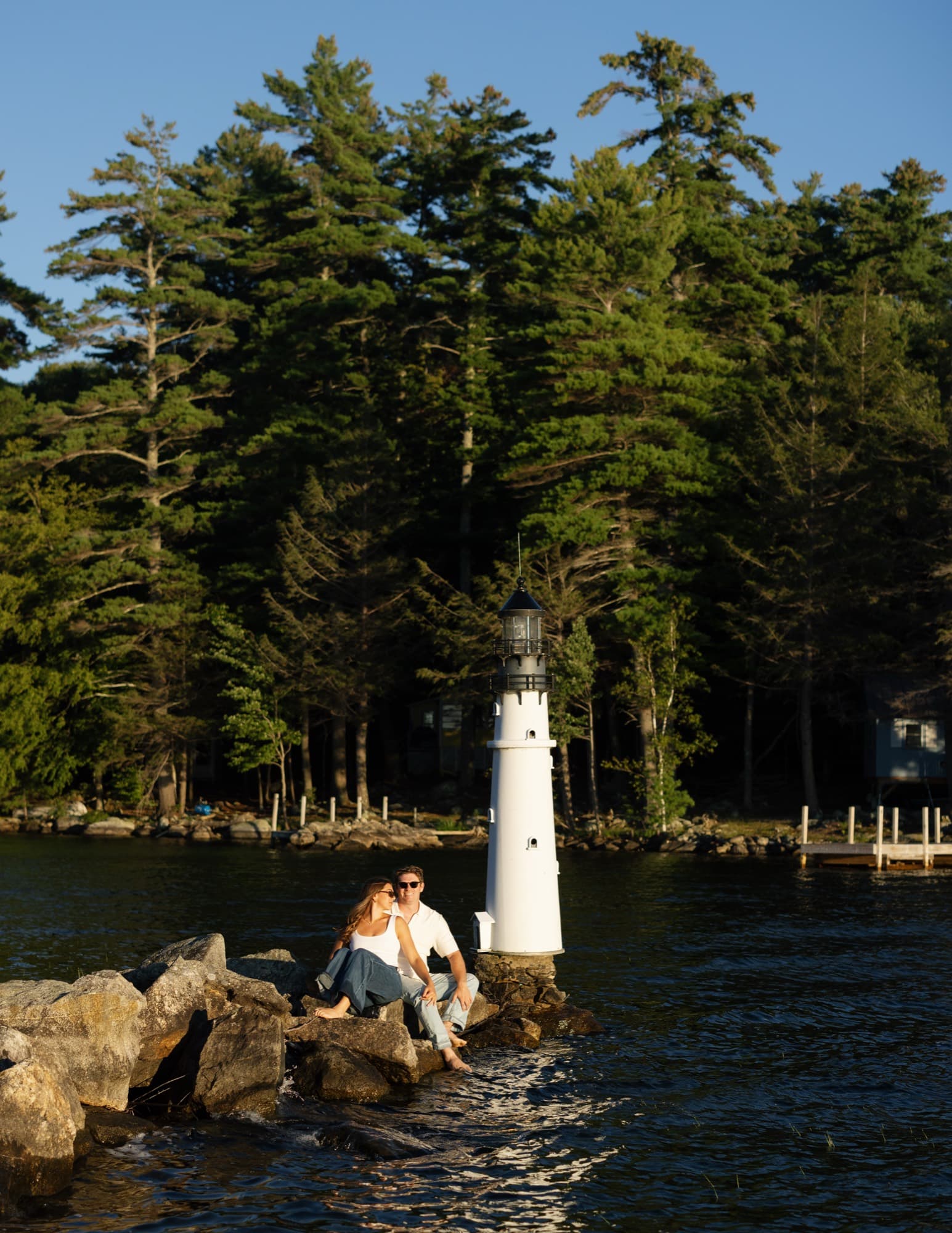 Morgan and Jon sitting together on rocks beside a small white lighthouse, tall pines rising behind.
