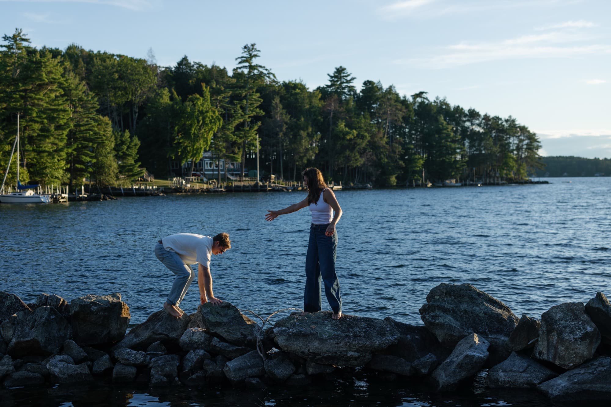 Morgan standing on a rock jetty reaching a hand back toward Jon as he climbs up, the wide lake and pine shoreline behind them.